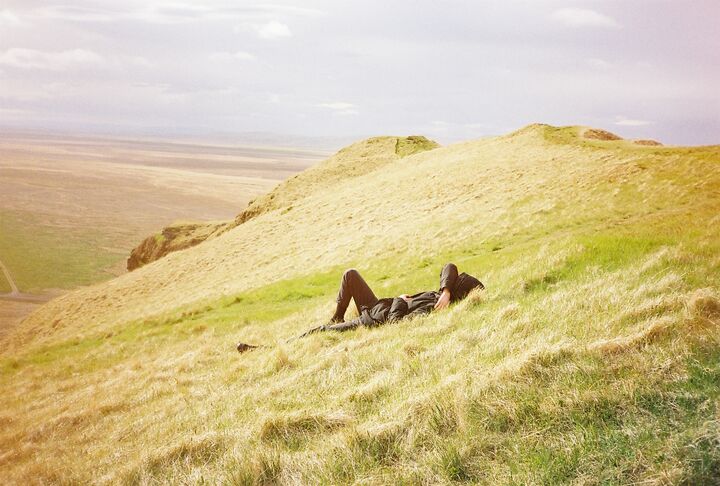 A person lying back in tall golden grass on a hillside, one arm resting over their face, with a hazy valley stretching out below.